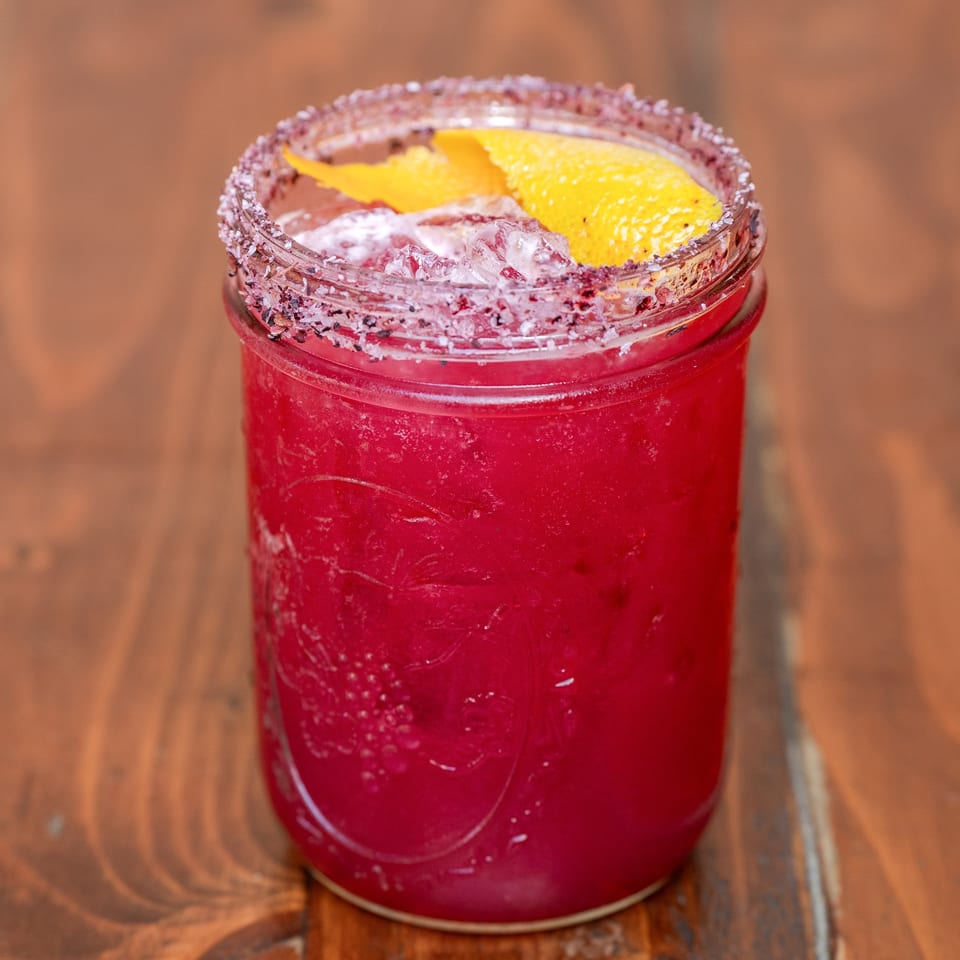 Que Pasa Cantina's vibrant red margarita in a mason jar, garnished with citrus peel and a salted rim.