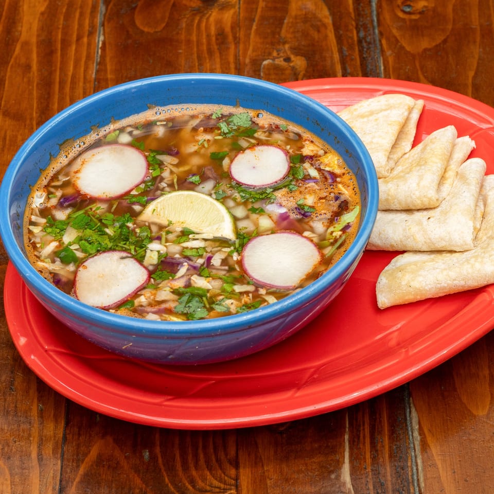 Que Pasa Cantina's pozole soup with radishes, lime, and cilantro, served with tortillas on a red plate.