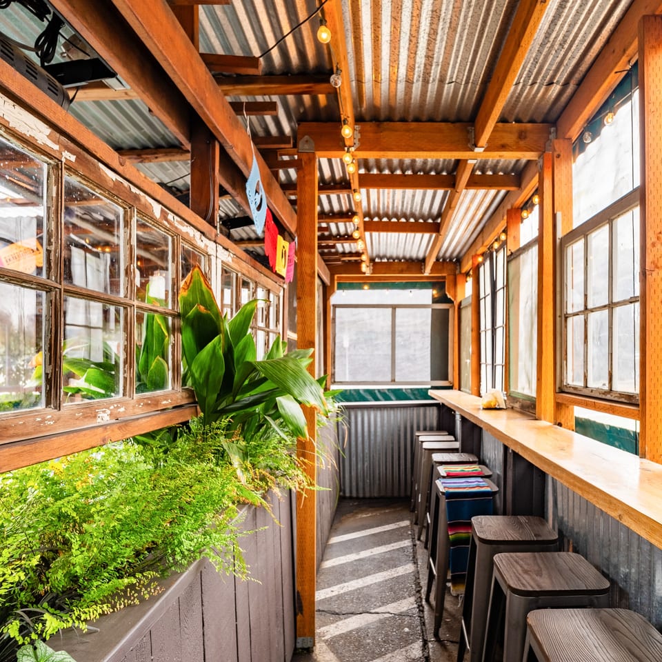 Que Pasa Cantina's outdoor patio with a bar, stools, plants, and string lights under a corrugated roof.