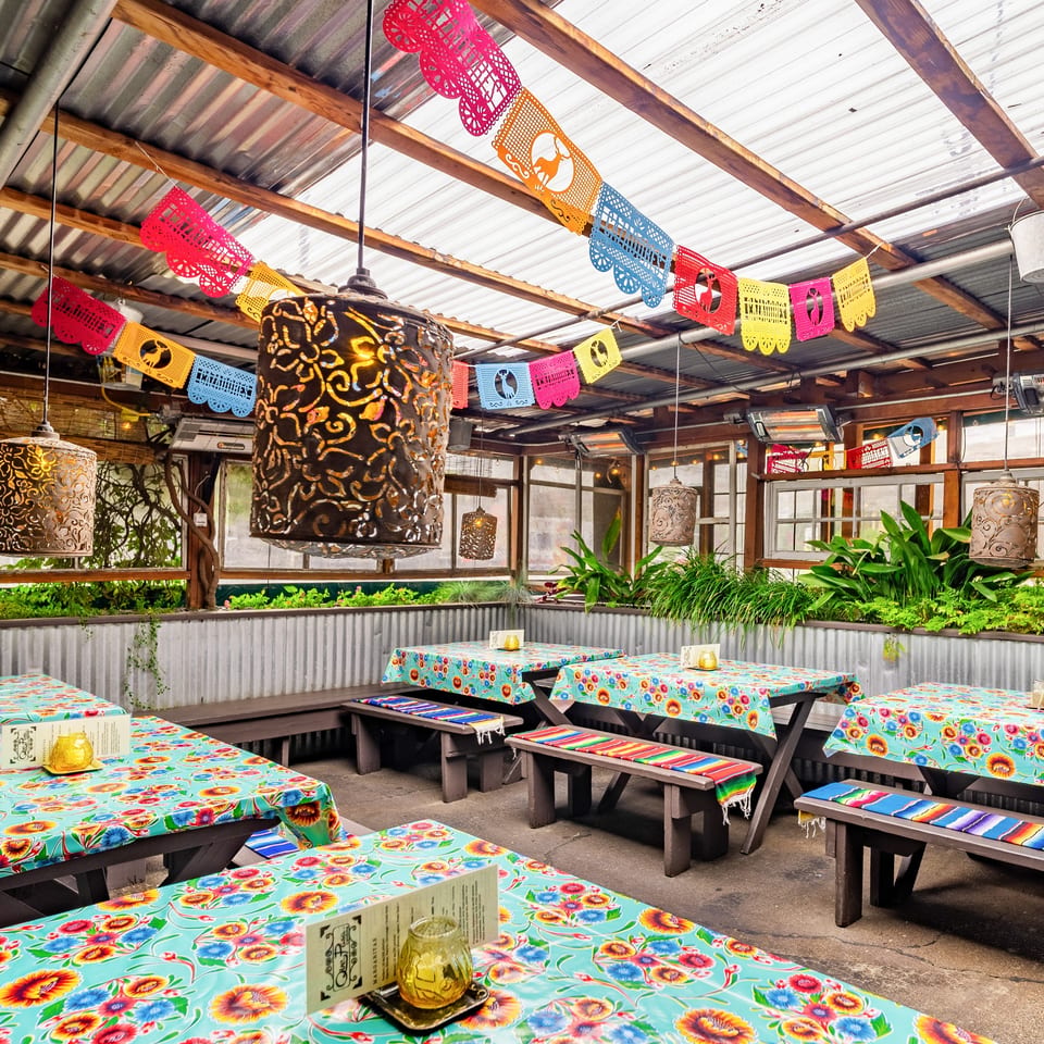 Que Pasa Cantina's vibrant outdoor patio with colorful papel picado, floral tablecloths, and rustic wooden benches.